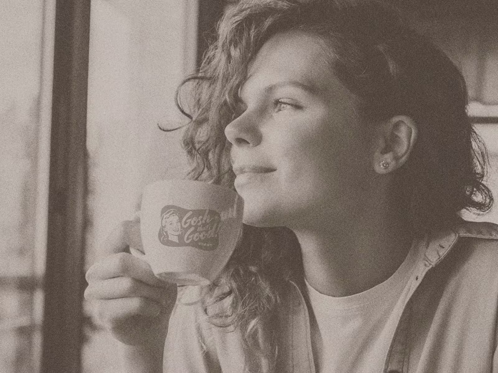 Sepia-toned photo of a woman holding a Gosh That's Good! branded mug while looking out a window.